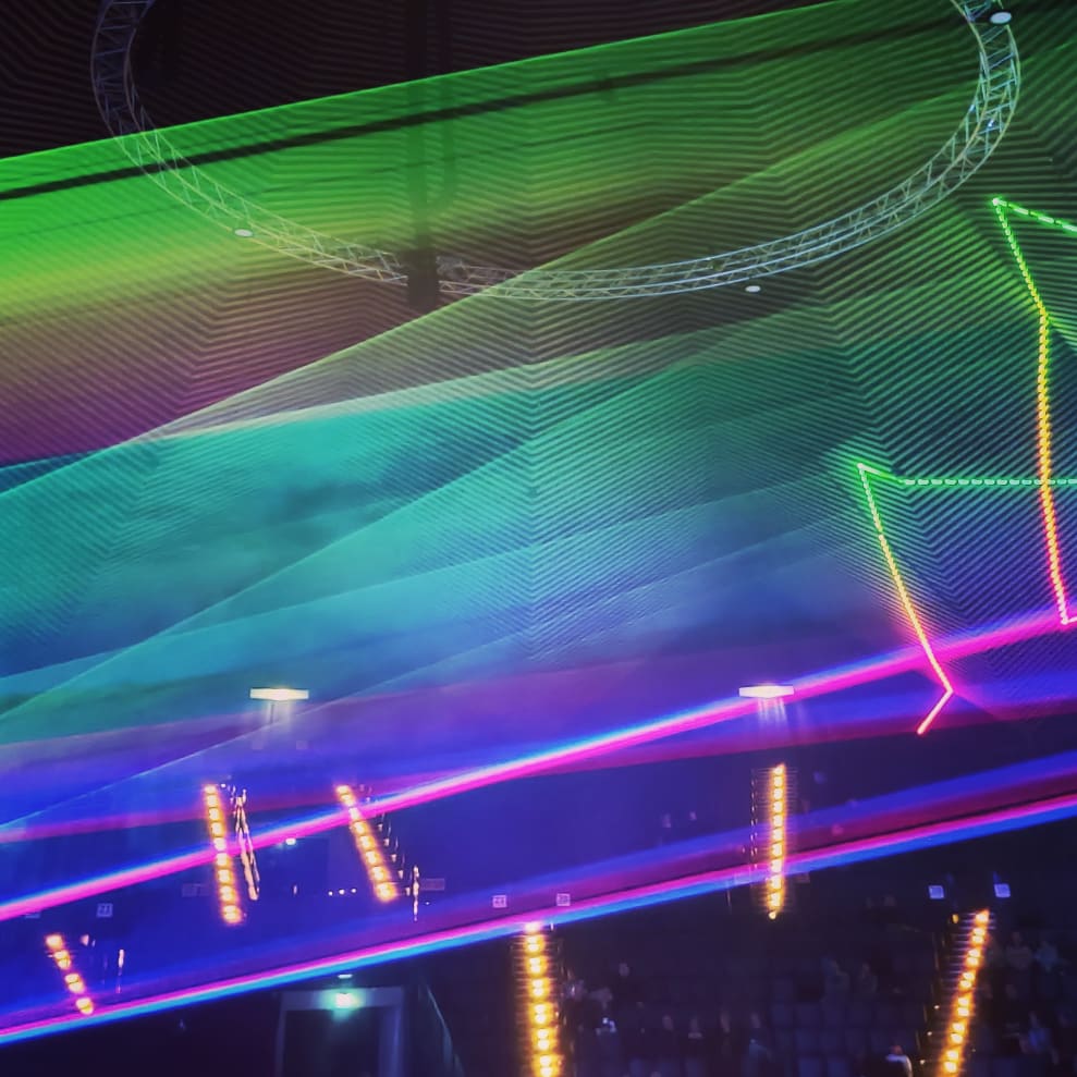 photo of a lasershow display during a concert. in the background, a crowd of people is visible, the laser show being a big green to blue gradient with some magenta stripes at the bottom